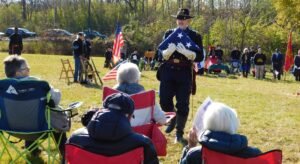 A man in a Civil War-era uniform holds a folded US flag and approaches a group of seated people