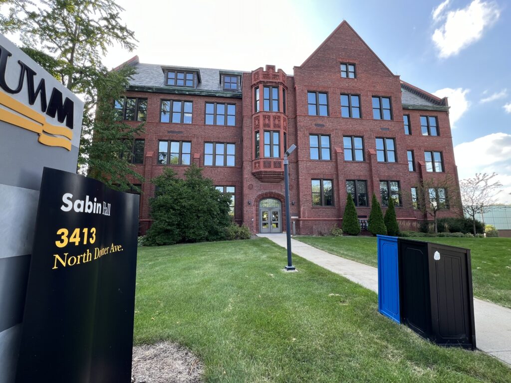 Photograph of a multi-story red brick building. A sign beside the walkway says, "UWM, Sabin Hall, 3413 North Downer Ave."