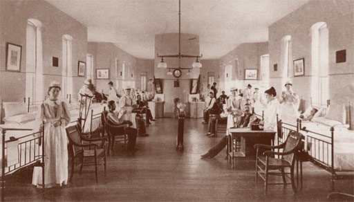 Historical photograph of a hospital ward with patients seated and lying in beds along both walls, attended by nurses. The room is long with high ceilings and large windows, evoking early institutional care settings of the late 19th or early 20th century.