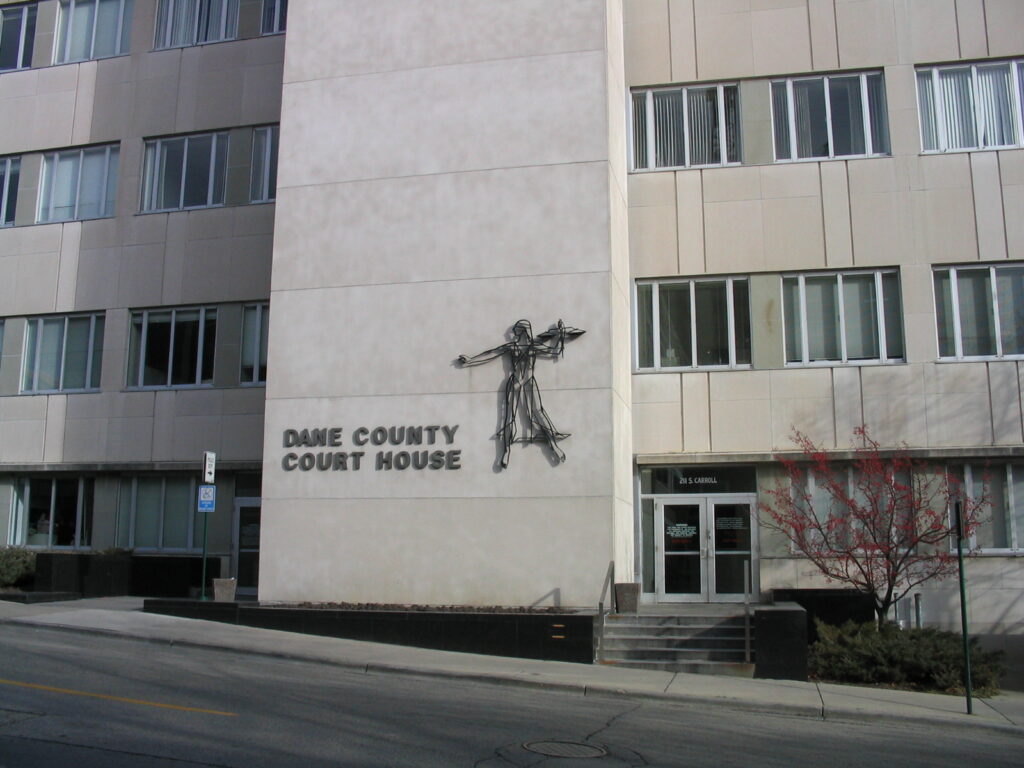 Dane County Courthouse. A sign identifies the building. There is a wireframe sculpture of a blindfolded Lady Justice holding scales.
